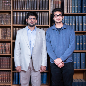 Two young men of South Asian descent. The man on the left is in a white two-piece suit and has a short beard. The man on the left is in a blue 3/4 zip jumper. In the background is a wall of books. 
